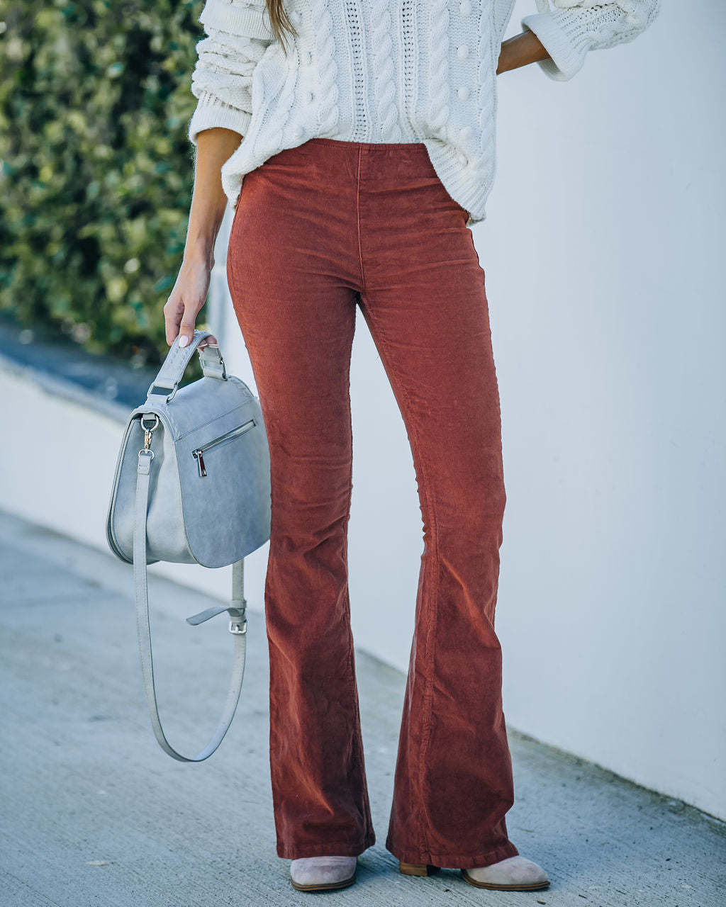 Femme en pantalon en velours côtelé marron, pull blanc torsadé, sac à main gris, posant à l'extérieur. Mode automne, style décontracté chic.