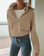 Femme portant un pull beige à col zippé, sur un balcon avec vue extérieure, combiné avec un jean bleu. Mode automne, style décontracté.