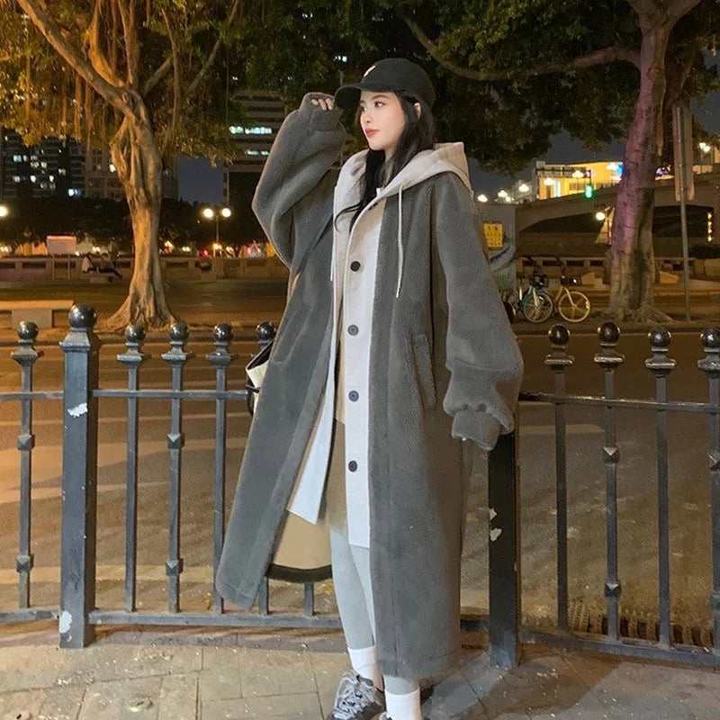 Jeune femme en manteau long gris, capuche blanche, posant près d'une clôture en ville la nuit. Mode urbaine, style décontracté, hiver.