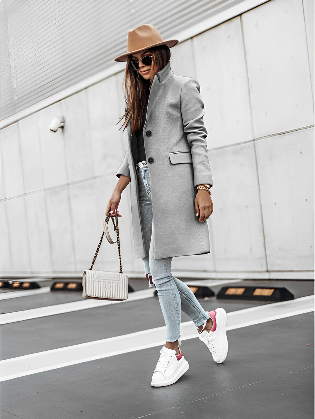 Femme élégante en manteau gris, chapeau marron, jeans déchiré et baskets blanches, portant un sac à main beige, posant devant un mur moderne.