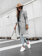 Femme élégante en manteau gris, chapeau marron, jeans déchiré et baskets blanches, portant un sac à main beige, posant devant un mur moderne.
