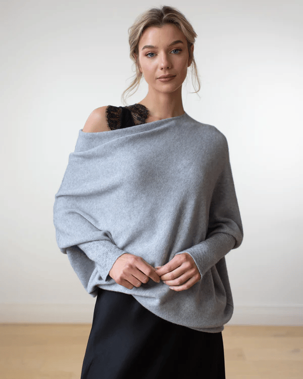 Femme portant un pull gris oversize élégant, avec une épaule dénudée, sur une robe noire. Mode automne-hiver, style décontracté chic.