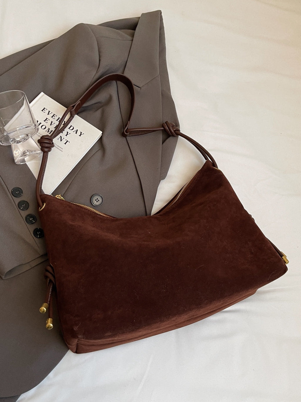 Sac à main en daim marron sur un manteau gris, avec un livre et un verre à côté. Mode élégante, accessoires tendance, style automne-hiver.