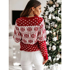 Femme portant un pull rouge à motifs flocons de neige blancs, debout devant un sapin de Noël décoré. Mode hiver, tenue festive, ambiance Noël.