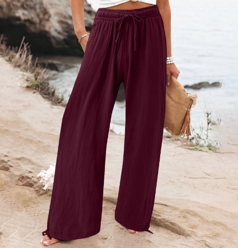 Pantalon large bordeaux femme, plage en arrière-plan, tenue estivale décontractée, accessoire sac paille, mode bord de mer.