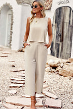 Femme en combinaison beige élégante, lunettes de soleil, bijoux dorés, marchant sur un chemin de pierres devant un bâtiment blanc. Mode estivale chic.