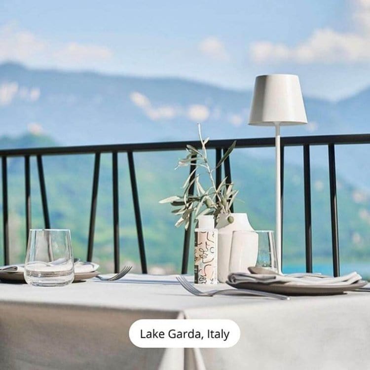 Table élégante avec vue sur le lac de Garde, Italie, comprenant des verres, couverts, lampe moderne et vase avec branches d'olivier.