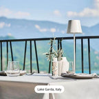 Table élégante avec vue sur le lac de Garde, Italie, comprenant des verres, couverts, lampe moderne et vase avec branches d'olivier.