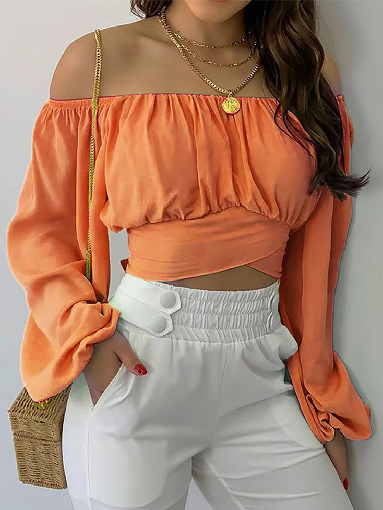 Femme portant un haut orange à épaules dénudées et un pantalon blanc, accessoirisée d'un collier doré et d'un sac en paille. Mode estivale élégante.