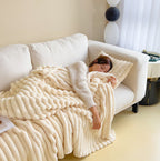 Femme endormie sur un canapé blanc, enveloppée dans une couverture en peluche beige. Intérieur cosy, ambiance détente, lumière naturelle.