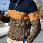 Homme portant un pull en tricot à rayures bleu marine, orange et marron, tenant des lunettes de soleil. Mode automne-hiver, style décontracté.