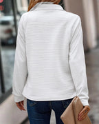 Femme portant un pull blanc texturé, manches longues, avec un sac à main beige, vue de dos, dans une rue urbaine. Mode automne-hiver élégante.