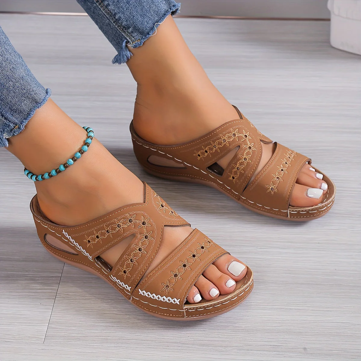 Sandales marron en cuir pour femme avec motifs brodés, portées avec un jean et un bracelet turquoise. Chaussures d'été confortables et élégantes.