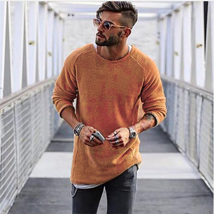 Homme moderne portant un pull orange, lunettes de soleil et accessoires, marchant sur une passerelle urbaine. Mode masculine, style décontracté.