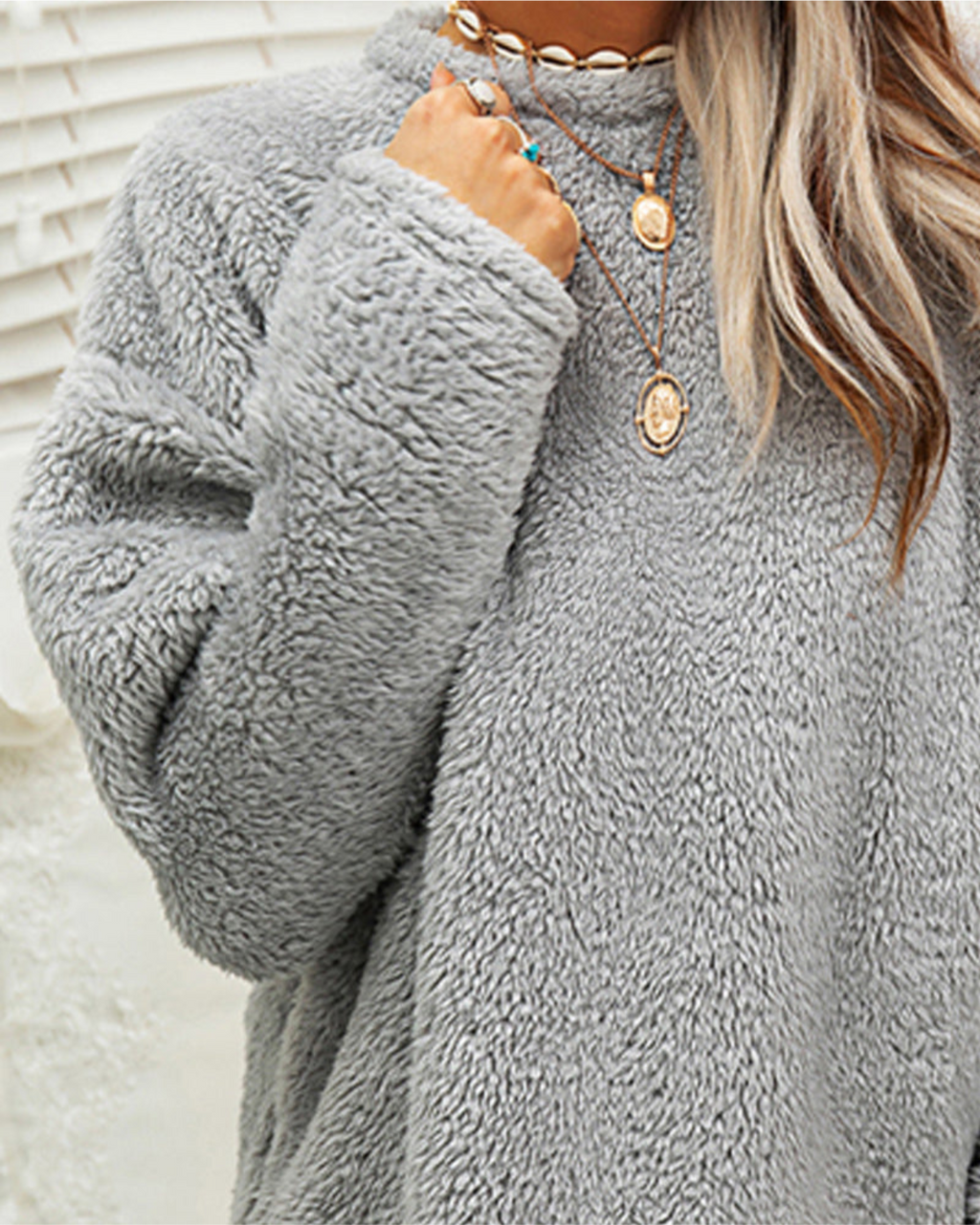 Femme portant un pull en fausse fourrure gris, col rond, avec colliers dorés superposés. Mode hiver, tendance cosy, style décontracté.