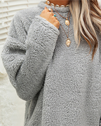 Femme portant un pull en fausse fourrure gris, col rond, avec colliers dorés superposés. Mode hiver, tendance cosy, style décontracté.