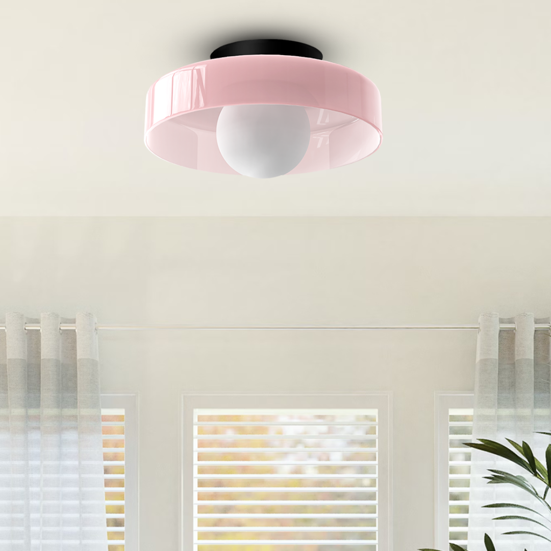 Plafonnier moderne rose avec abat-jour circulaire, installé au plafond d'un salon lumineux avec rideaux gris et fenêtres à persiennes blanches.