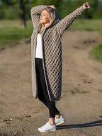 Femme portant un long cardigan en tricot beige avec capuche, debout sur un chemin de terre, bras levés, style décontracté, mode automne.