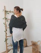 Femme portant un châle en tricot gris, debout près d'une échelle en bois décorée de feuillage, dans un intérieur moderne et minimaliste.