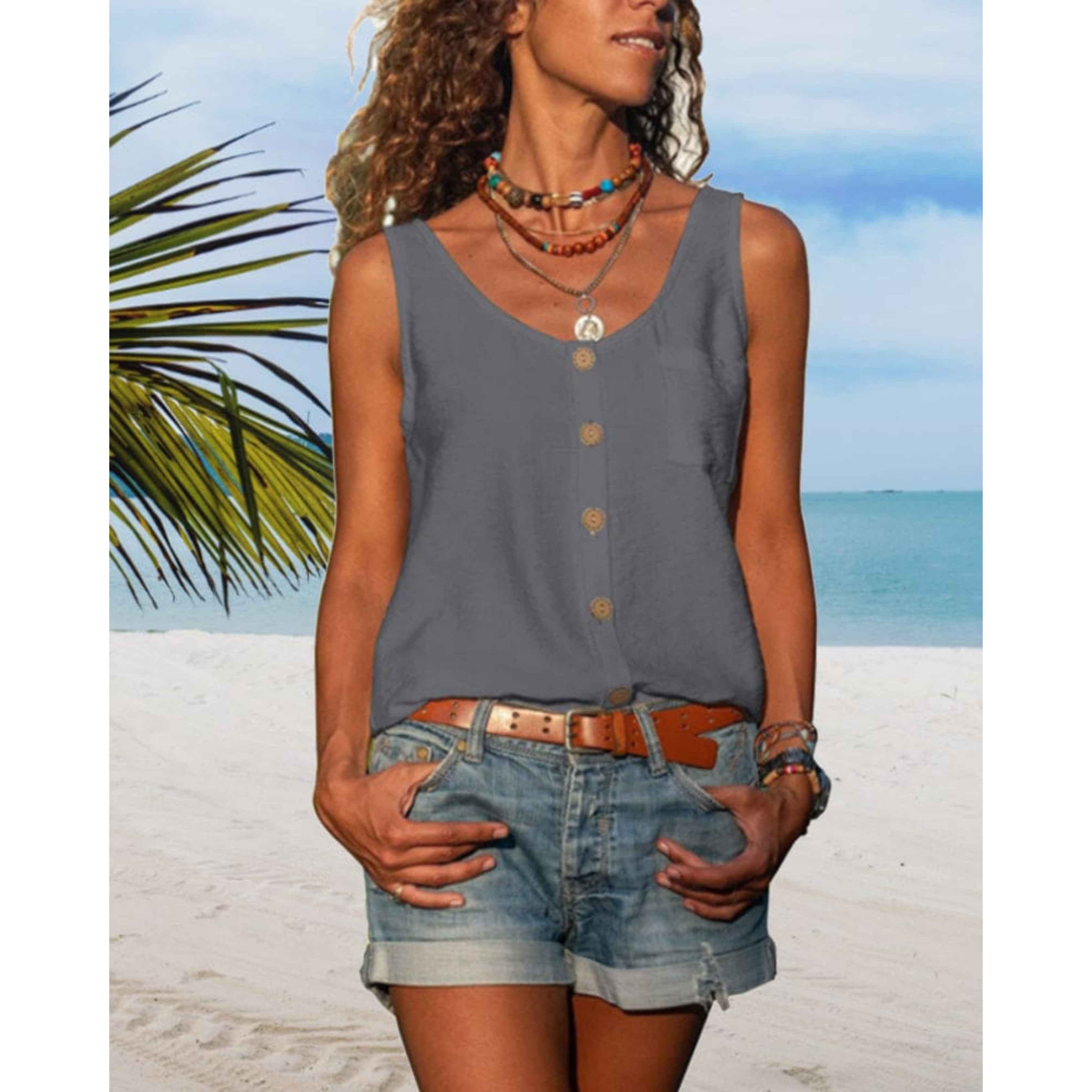 Femme en débardeur gris et short en jean sur plage tropicale, collier coloré, ciel bleu, mer en arrière-plan, ambiance estivale et décontractée.