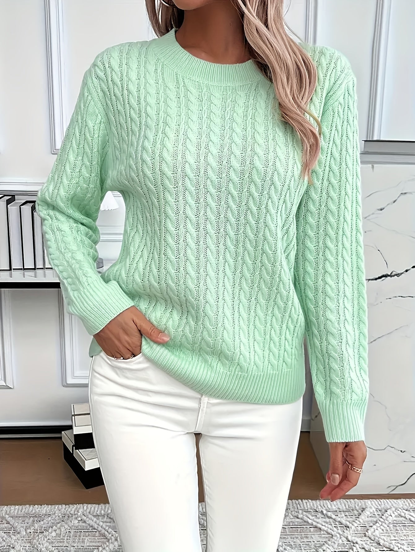 Femme portant un pull torsadé vert menthe et un pantalon blanc, posant dans un intérieur moderne. Mode automne, style décontracté, tendance féminine.