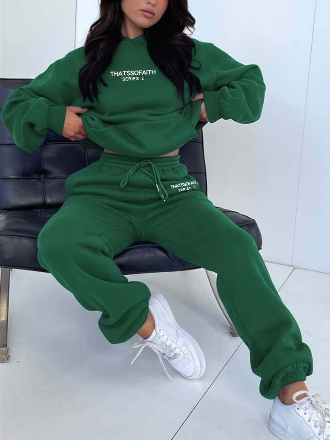 Femme en survêtement vert, assise sur un fauteuil noir, portant des baskets blanches. Mode décontractée, tendance streetwear, style confortable.