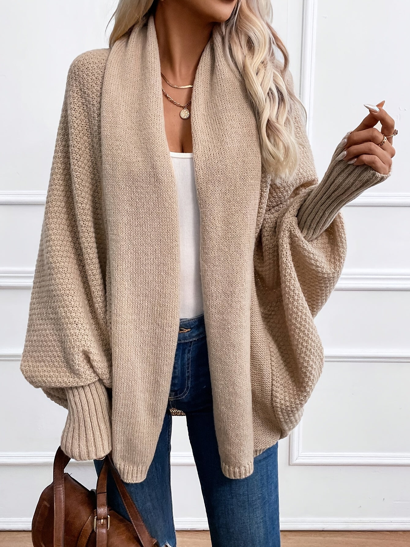 Femme portant un cardigan beige oversize en tricot, manches longues, avec un top blanc et un jean bleu, accessoirisé de colliers dorés.
