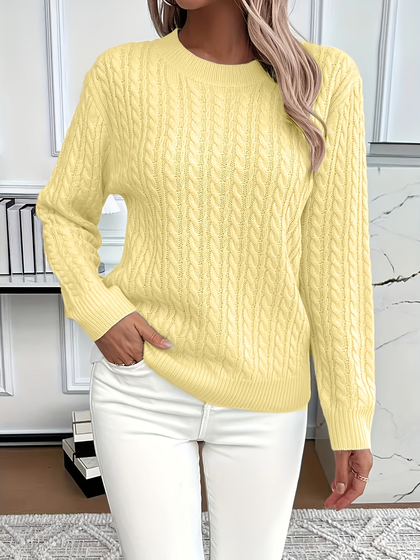 Femme portant un pull jaune en maille torsadée, manches longues, avec un pantalon blanc. Mode automne, style décontracté, intérieur moderne.