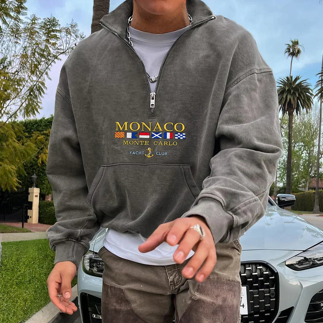 Homme portant un sweat-shirt gris "Monaco Monte Carlo Yacht Club" devant une voiture de luxe, entouré de palmiers. Mode décontractée, style urbain.
