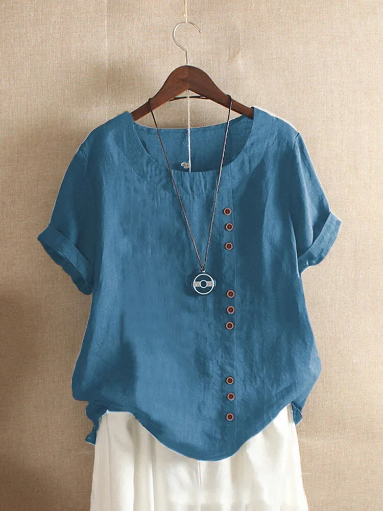 Blouse en lin bleu avec boutons asymétriques, manches courtes retroussées, col rond, suspendue sur cintre, fond beige. Mode femme élégante.