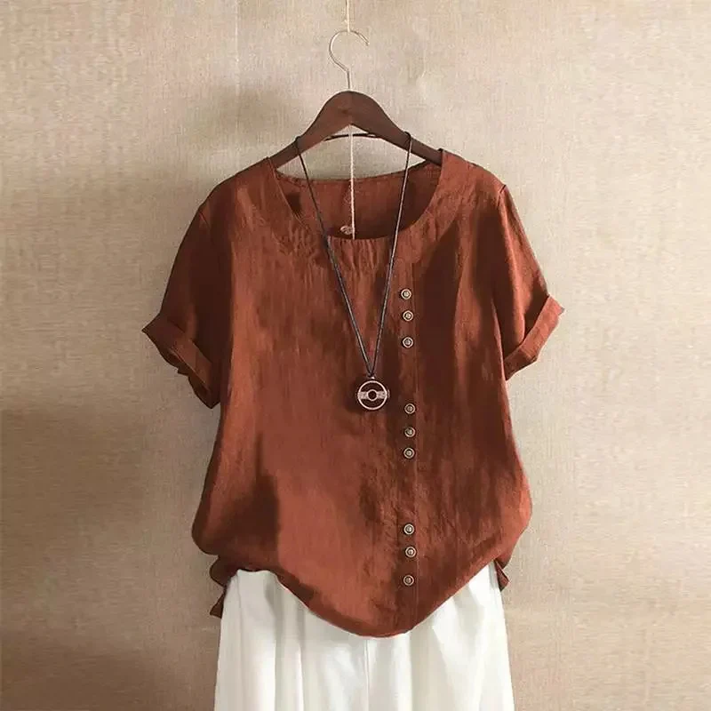Blouse en lin marron avec boutons asymétriques, manches courtes retroussées, suspendue sur un cintre, collier pendentif rond, fond beige.