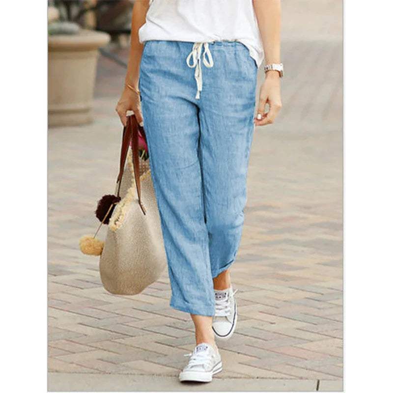 Femme portant un pantalon en lin bleu clair, baskets blanches et un sac en toile beige, marchant sur un trottoir pavé. Mode décontractée estivale.