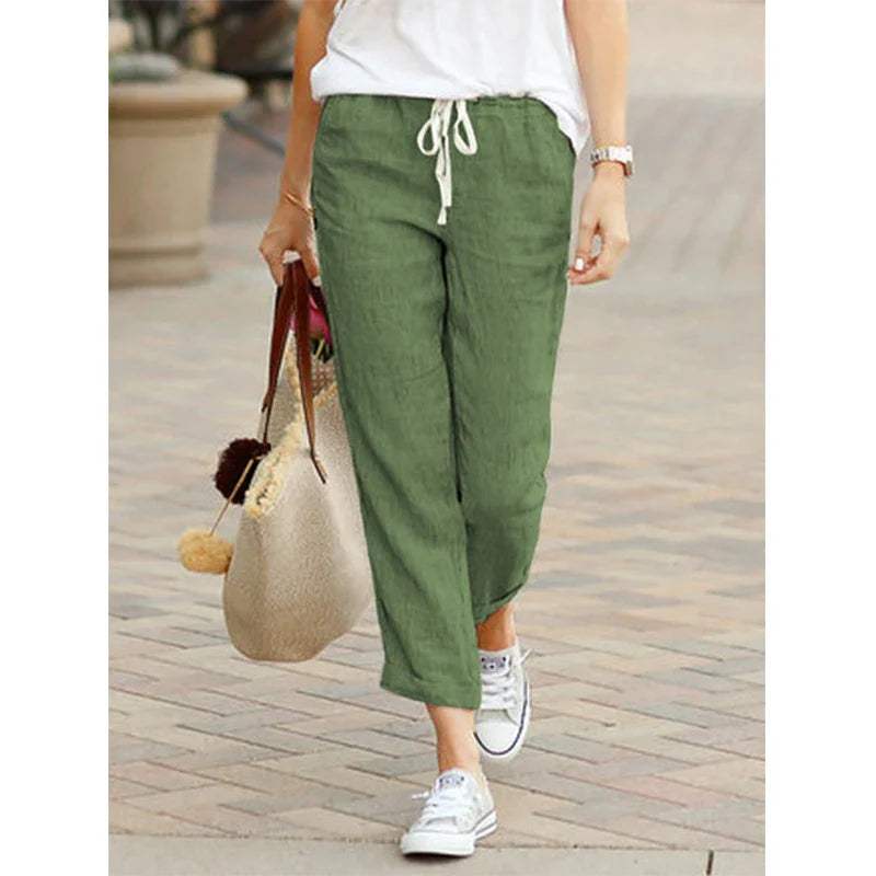 Femme portant un pantalon vert en lin, t-shirt blanc, baskets blanches, sac en paille; mode décontractée, tenue estivale élégante.