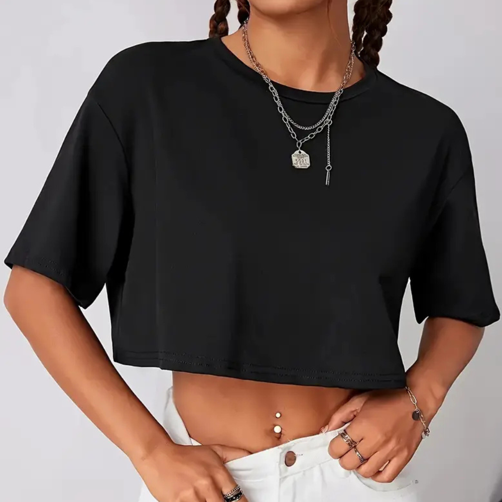 Femme portant un crop top noir, col rond, manches courtes, avec un collier en argent, posant en jean blanc. Mode tendance, style décontracté.