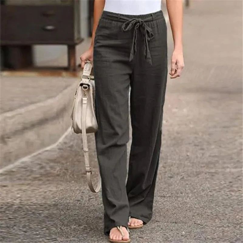 Pantalon large noir femme avec ceinture nouée, porté avec un haut blanc et des sandales, sac à main beige, mode décontractée, rue pavée.