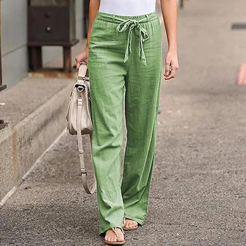Femme portant un pantalon vert en lin avec un haut blanc, sandales et sac à main beige. Mode décontractée, tenue estivale élégante en ville.