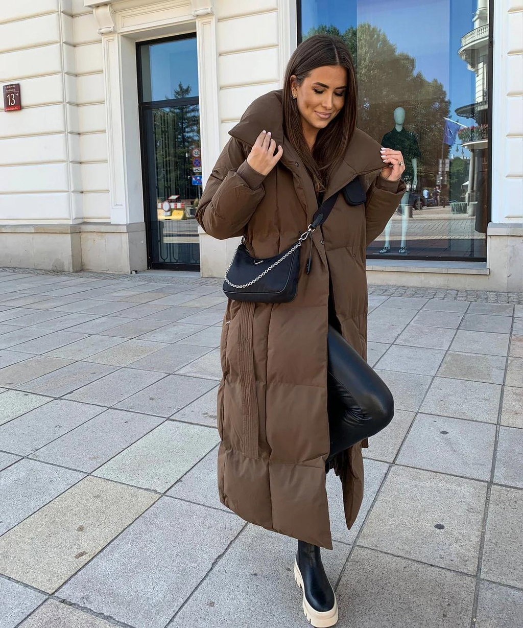 Femme portant un long manteau marron matelassé, sac à main noir, leggings en cuir, baskets blanches, devant un bâtiment moderne. Mode automne-hiver.