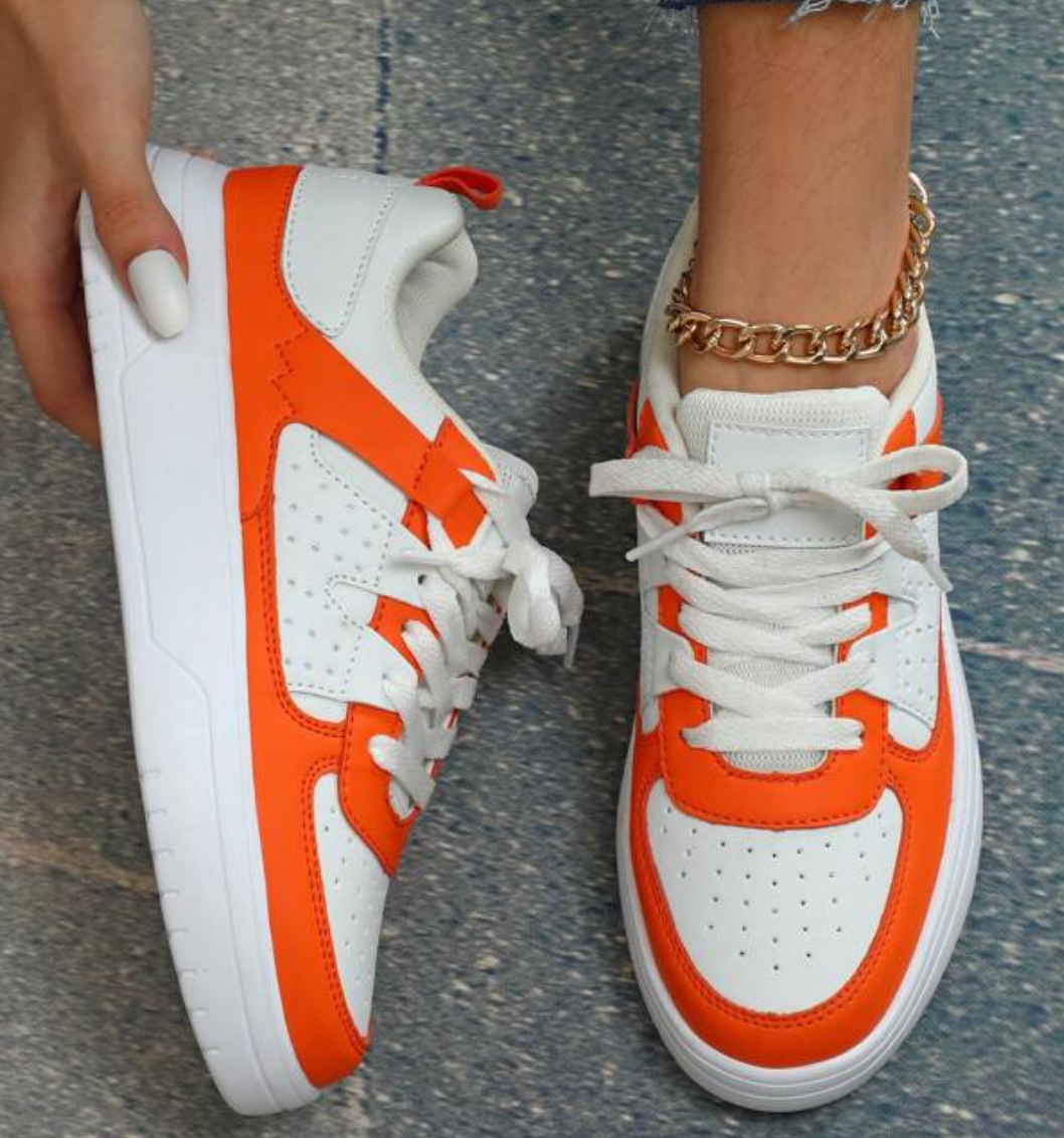 Chaussures de sport blanches et orange vif, design moderne, portées par une personne avec un bracelet doré, sur fond de sol en béton.