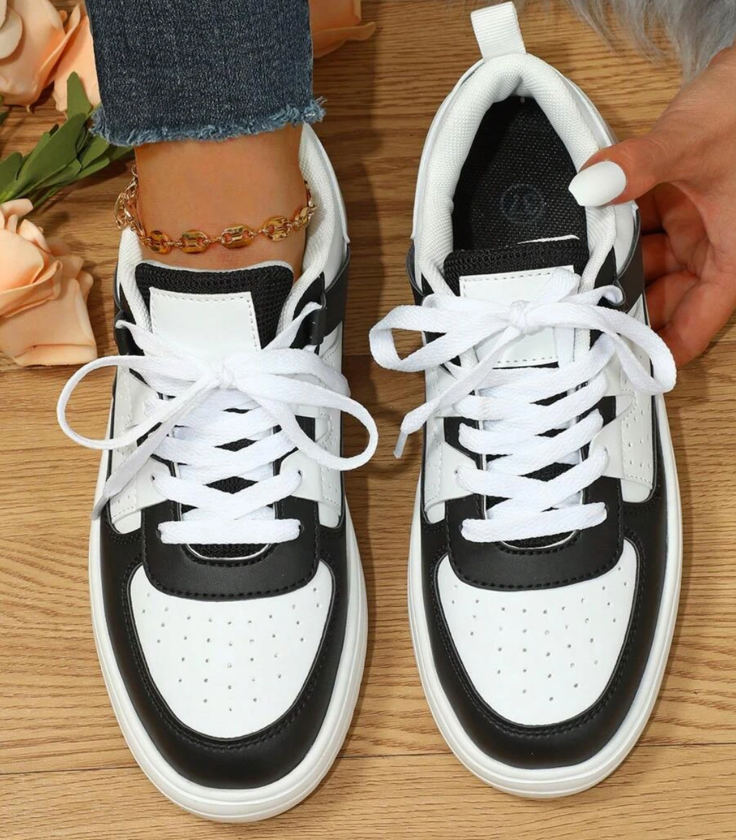 Chaussures de sport noires et blanches pour femme, lacets blancs, portées avec un jean et un bracelet de cheville doré, posées sur un sol en bois.