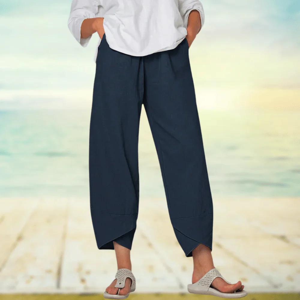Pantalon large bleu marine pour femme, style décontracté, porté avec un haut blanc et des sandales, sur fond de plage ensoleillée.