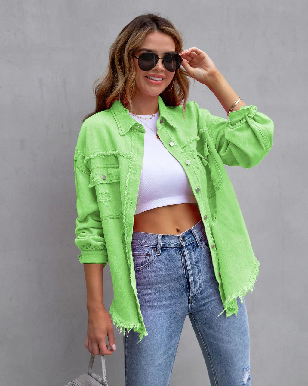 Jeune femme souriante portant une chemise verte fluo, un crop top blanc et un jean décontracté, posant contre un mur gris avec des lunettes de soleil.