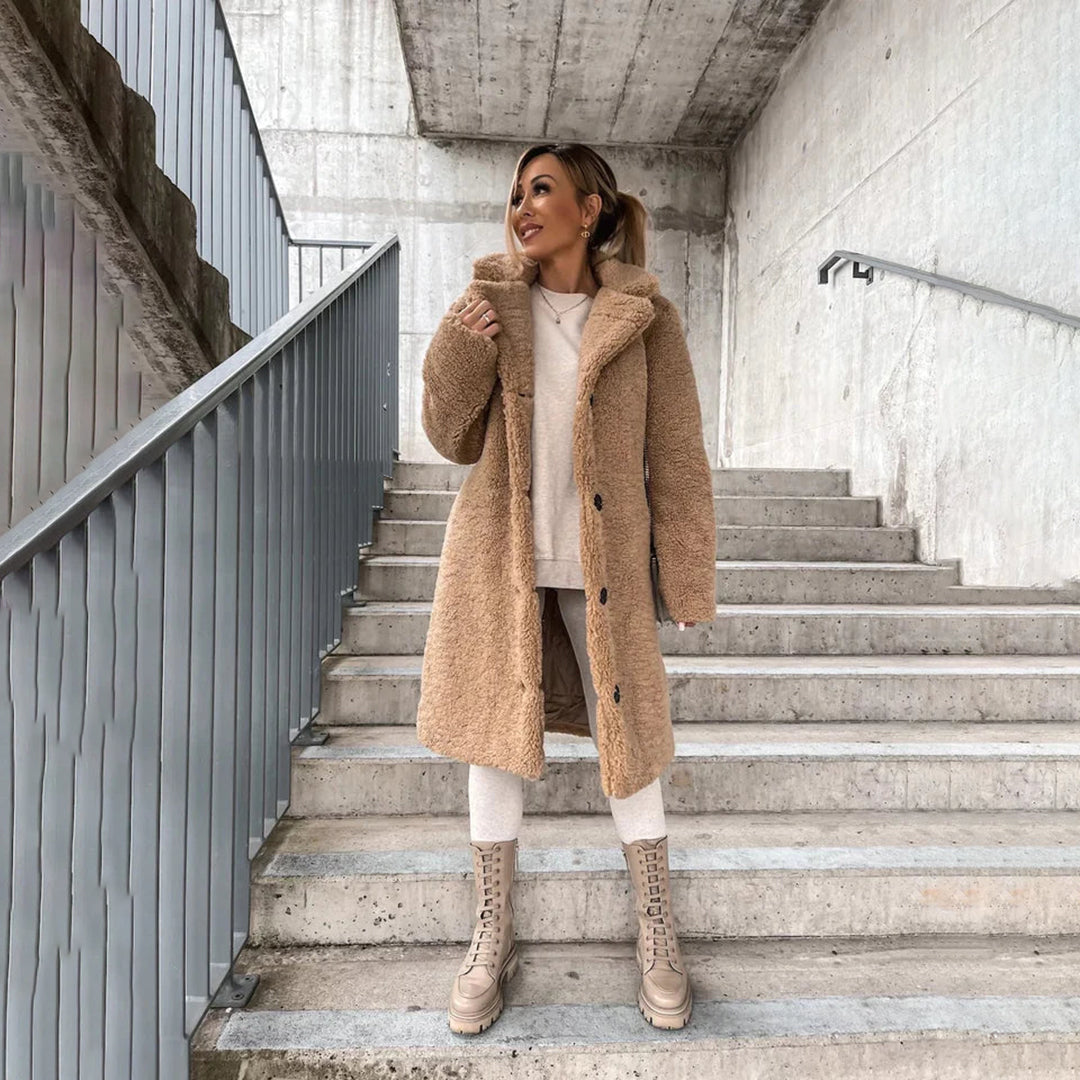 Femme portant un manteau en fausse fourrure beige, debout sur des escaliers en béton, mode hiver, style urbain, bottes beiges, look moderne.