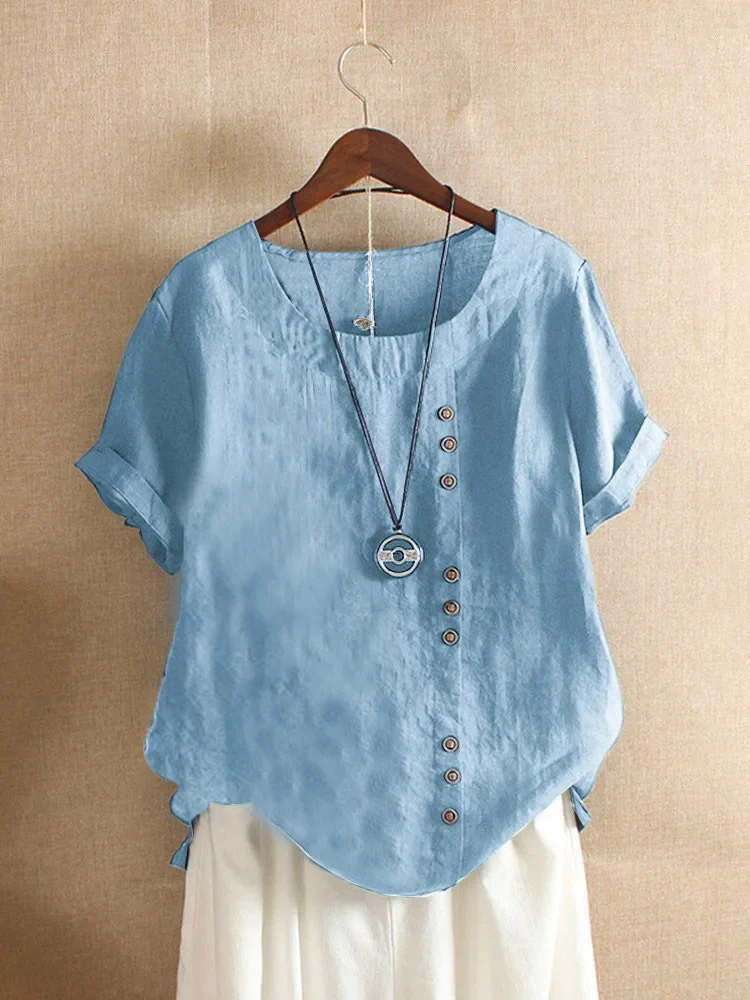 Blouse en lin bleu clair avec boutons asymétriques, manches courtes retroussées, col rond, suspendue sur un cintre, collier pendentif rond inclus.