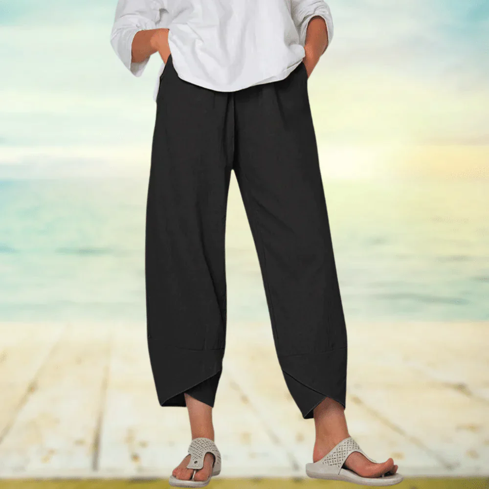 Pantalon large noir pour femme, style décontracté, avec ourlets asymétriques, porté avec un haut blanc et des sandales, sur fond de plage ensoleillée.