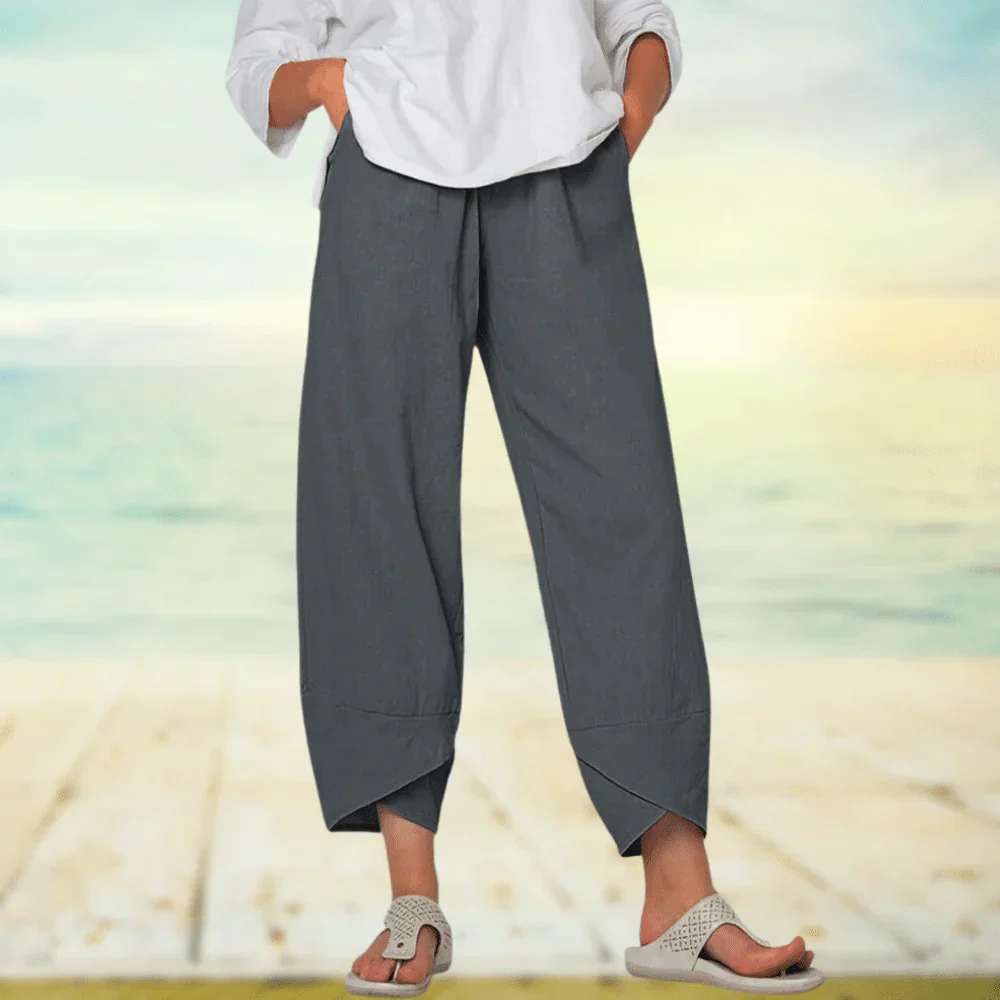 Pantalon ample gris pour femme, style décontracté, porté avec une chemise blanche et des sandales, sur fond de plage ensoleillée.