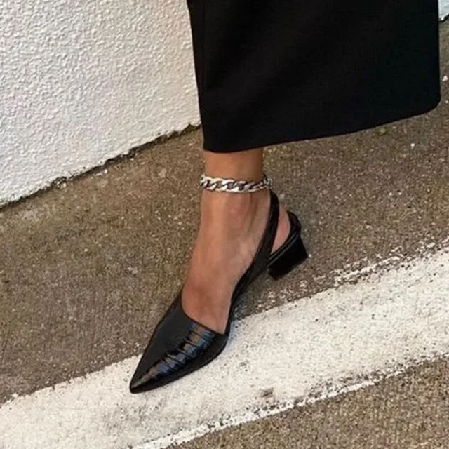 Escarpin noir à talon bas sur pied féminin, bracelet chaîne argentée, jupe noire, sol béton. Mode élégante, chaussures tendance, style urbain.