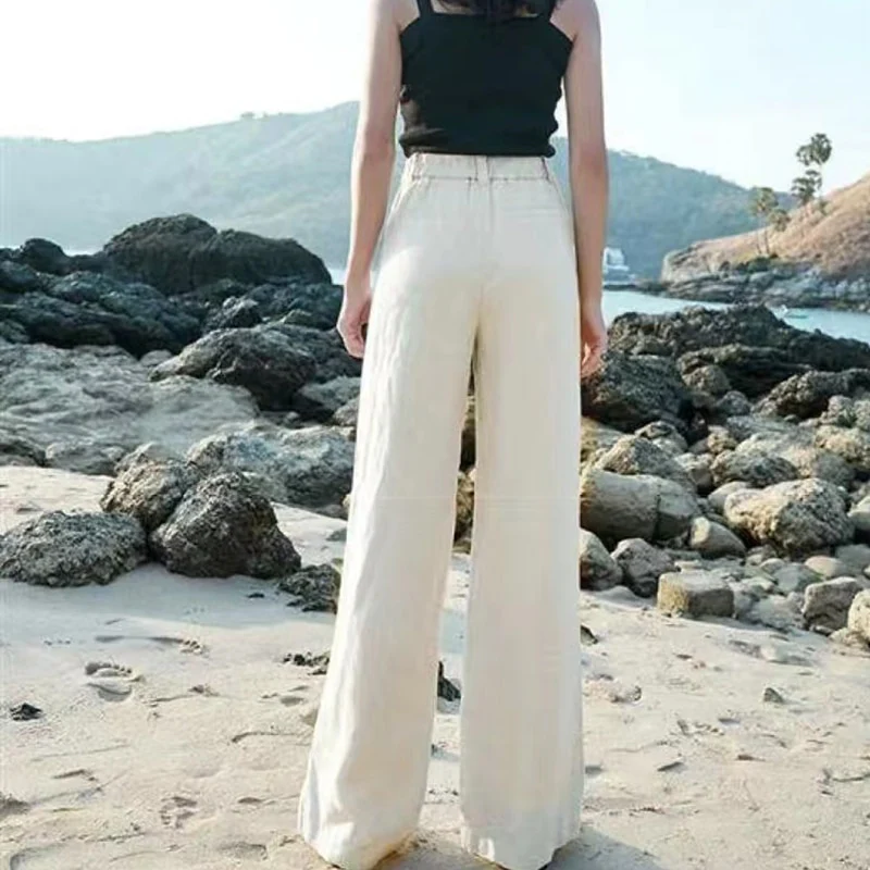 Femme en pantalon large blanc sur plage rocheuse, vue de dos. Mode estivale, tenue décontractée, paysage côtier.