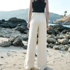 Femme en pantalon large blanc sur plage rocheuse, vue de dos. Mode estivale, tenue décontractée, paysage côtier.