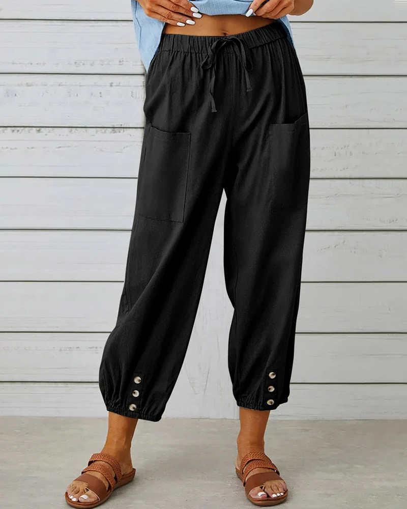 Pantalon noir ample avec poches avant, boutons décoratifs aux chevilles, porté par une personne debout sur fond de planches en bois.
