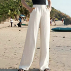 Personne portant un pantalon large blanc sur une plage ensoleillée, avec des palmiers en arrière-plan. Mode estivale, style décontracté, vacances.